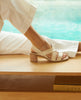 Woman sitting on wooden bench wearing Coclico Jade Heel in Greige leather: open slip on sandal with leather tube straps across the foot, sling-back strap, wood block mid-height heel. 7