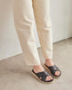 Woman modeling the Coclico Pasque Sandal in Black leather: low pine wedge slide sandal, rubber sole, intertwined leather tube straps across foot. 2
