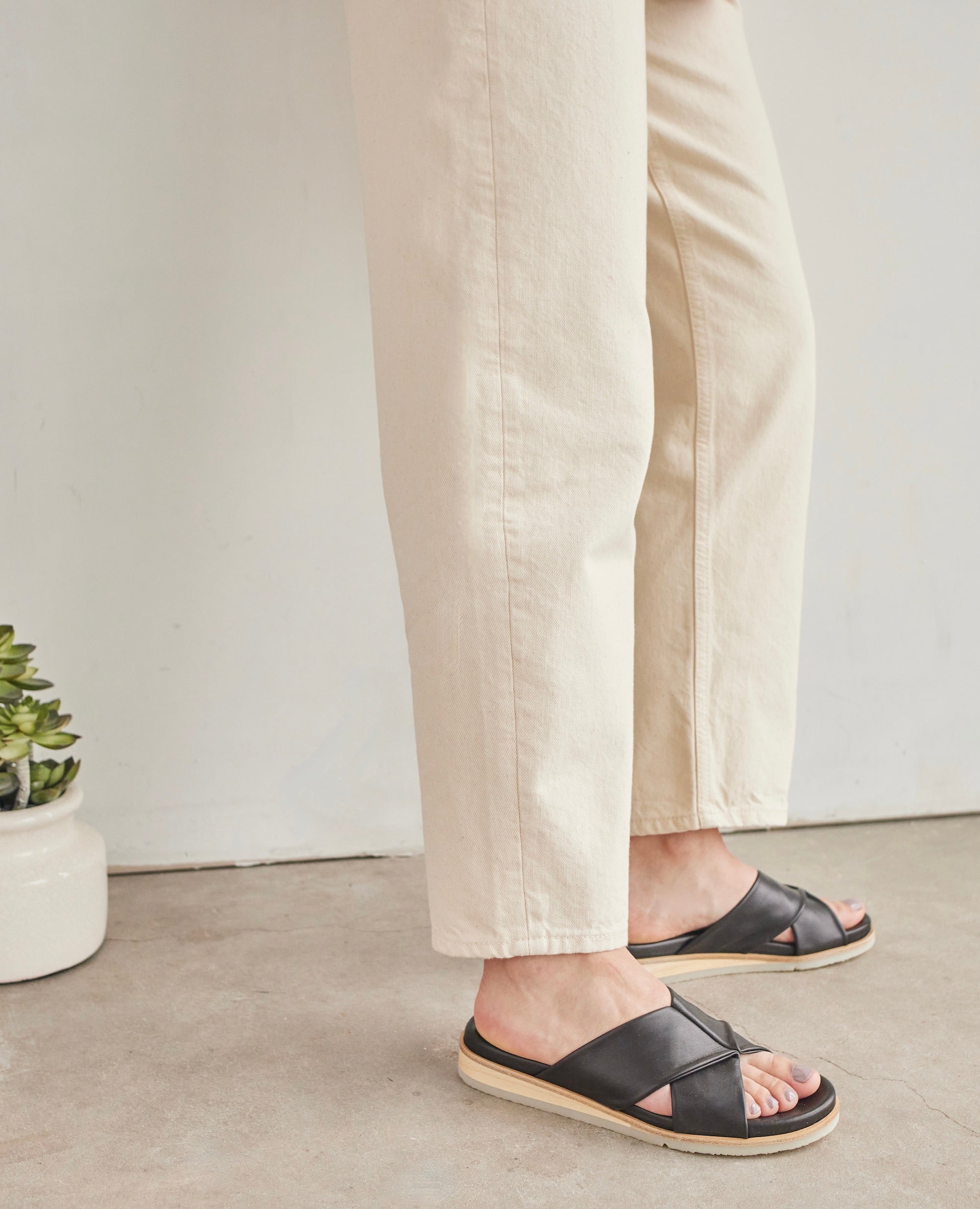Side view of woman modeling the Coclico Pasque Sandal in Black leather, a low pine wedge slide sandal with rubber sole and intertwined leather tube straps across foot.