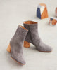 Close up of the perfect boot that will take you from day to night featuring a seam ankle detailing, mid-height solid wood block heel and an inside zip closure - Coclico Pimiento Boot in Anthracite suede with wooden art decor in background. 3
