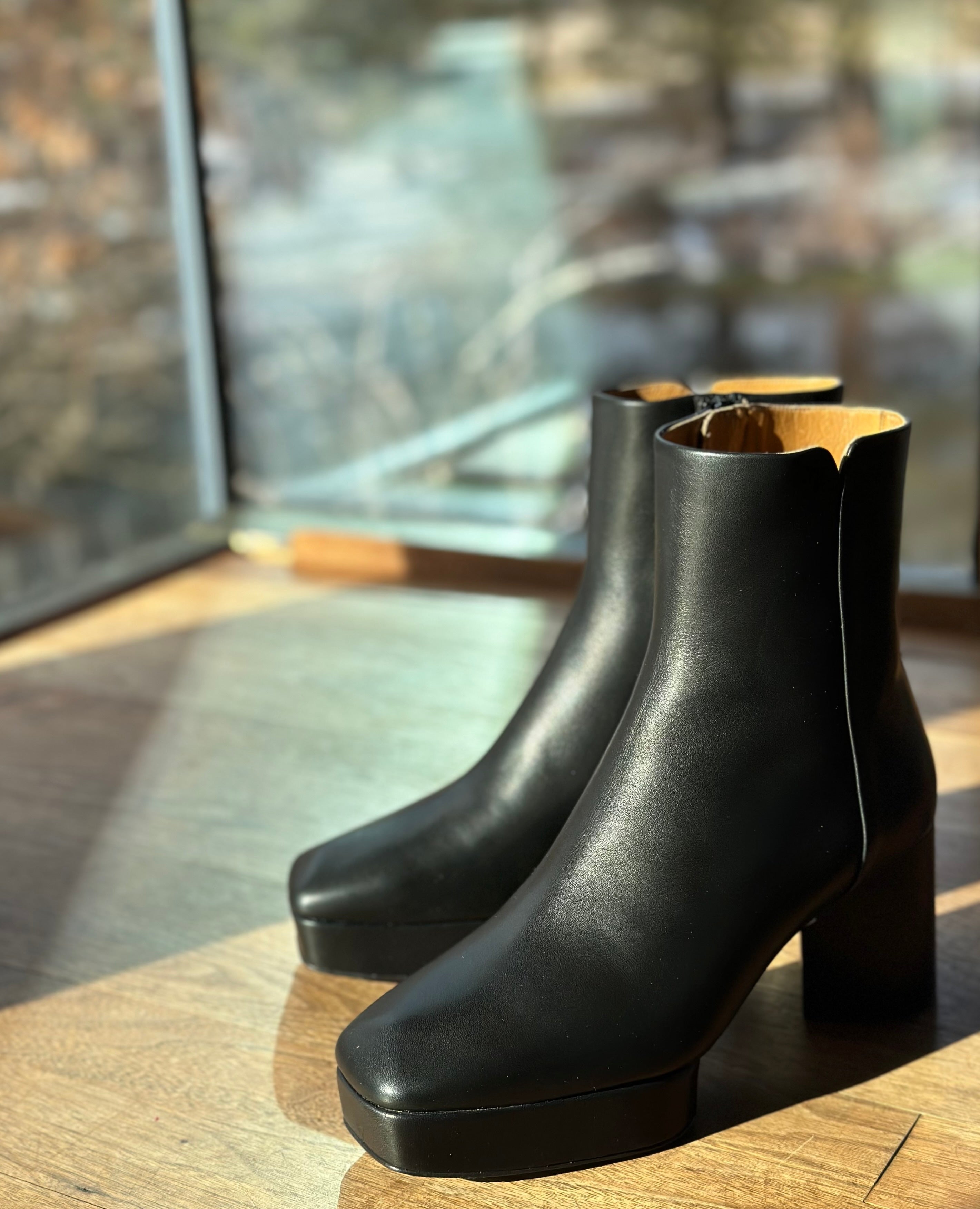 The Travis Boot in Black placed on a wooden floor with a window in the background and the sun shining.