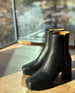 The Travis Boot in Black placed on a wooden floor with a window in the background and the sun shining.  4