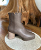 Side view of the Travis Boot in Taupe on a white rug with wood in the back. 5