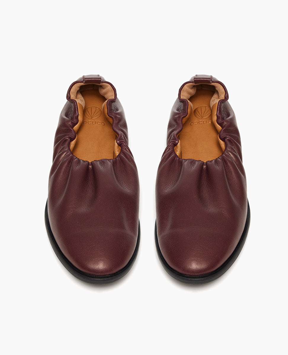 A pair of burgundy leather slip-on shoes with a gathered, ruched design around the opening, featuring a smooth, rounded toe and a black rubber sole.