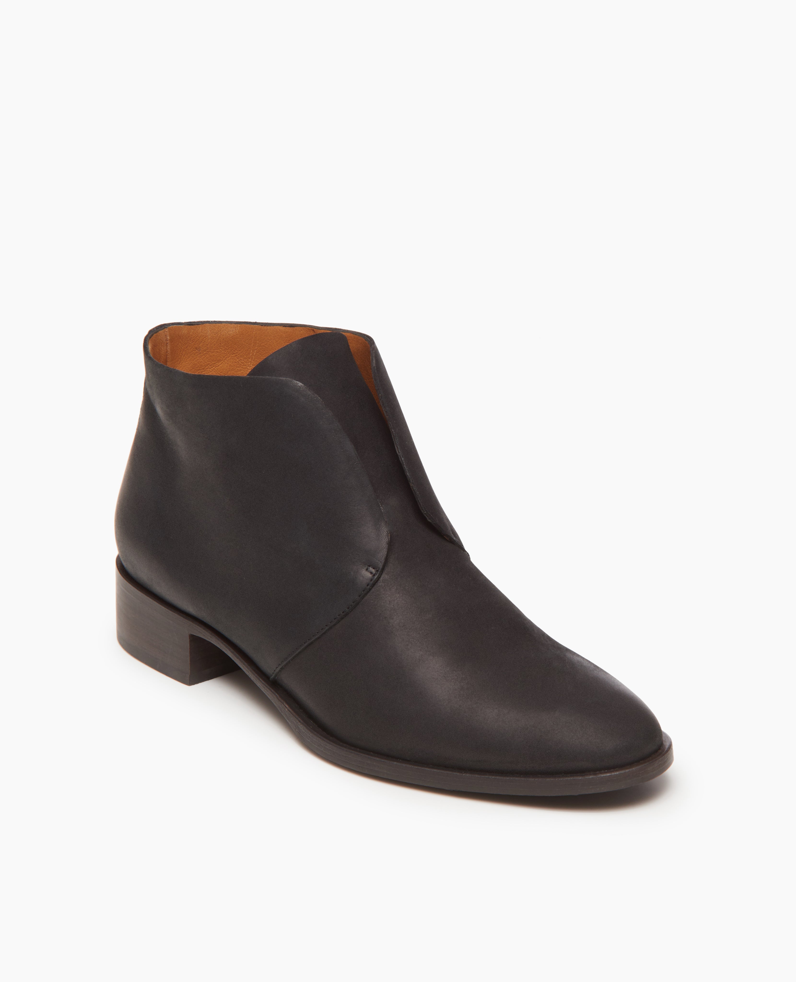 A sleek, black leather ankle boot with a smooth texture, minimalist design, and a low, stacked heel, featuring a unique asymmetrical opening and a tan interior lining.
