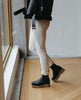 Woman modeling the Coclico Cappucho Boots in Black leather, a slip-on chelsea boot with water-resistant treated soles and a mid-height EVA sole platform - on wood flooring by large window. 5