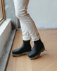 Model in Coclico Cappucho Boots in Black leather standing on wood flooring by a large window. 6