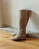 Coclico Chip Boot in Limestone nubuck, a knee-high boot with elastic goring, seam detailing across arch, inside zip closure, and a low-height solid wood block heel - leaning against white wall on wooden flooring. 2
