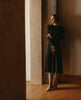 Model leaning of concrete wall wearing the Coclico Chip Boot in Limestone nubuck, a knee-high boot with elastic goring, seam detailing across arch, inside zip closure, and a low-height solid wood block heel - on wood flooring. 4