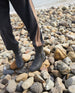 Individual in long black pants displaying Durum Shearling Boot on rocky beach. This black boot is displayed at an front angled view to show its full shearling interierior, outside zip enclosure, and updated upper lug sole.  7