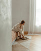 Model sitting on a bed putting on a slip-on, form-fitting flat in Italian certified sustainable leather with a squared-off toe and a 10mm solid wood heel - Coclico Haloumi Flat in Greige leather. 5