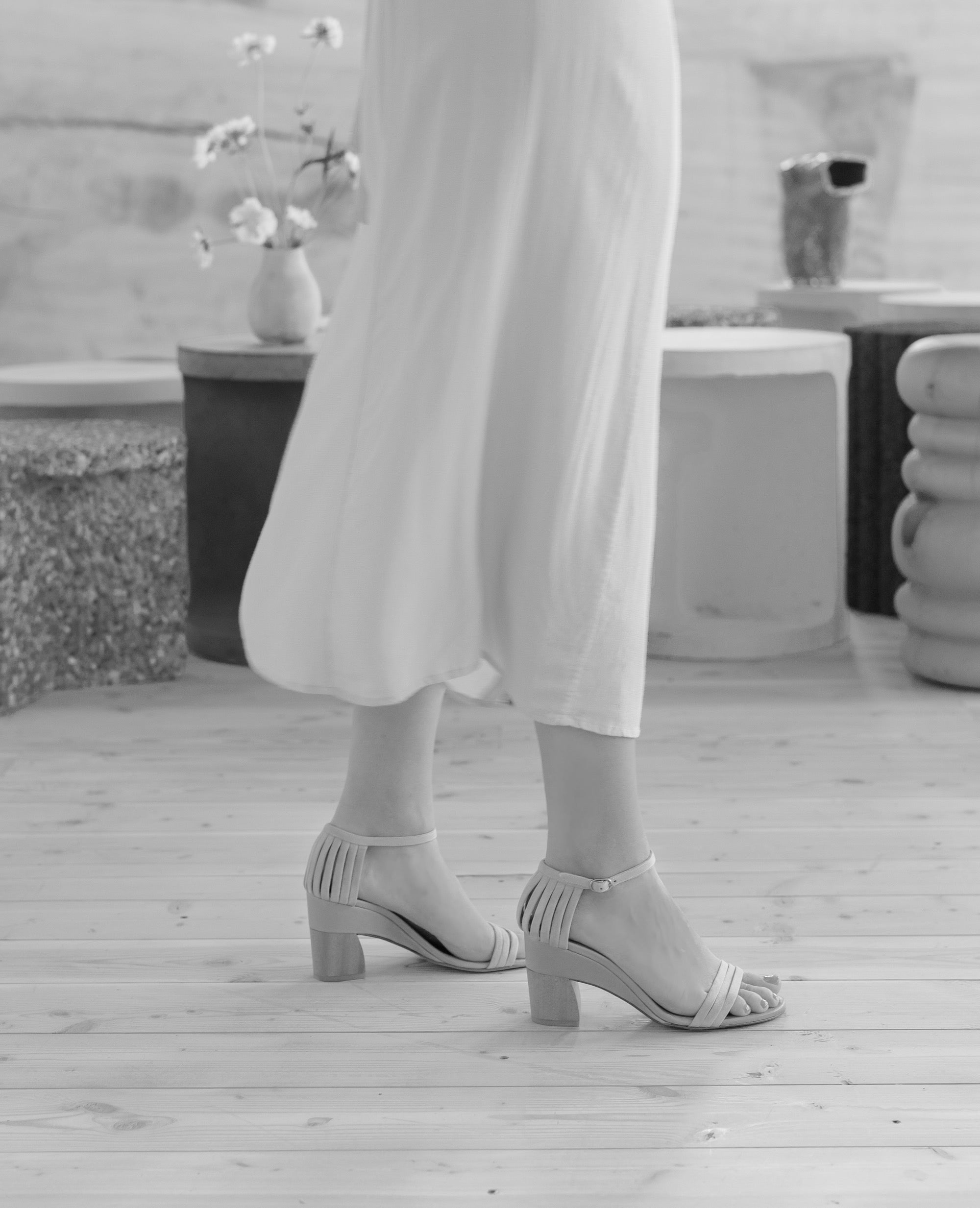 Woman modeling Coclico Iris Heel in Greige leather: 3 thin straps across front of the foot, fan effect of straps cupping heel, leather-wrapped demi-wedge, mid-height walnut wood heel - black & white side view.