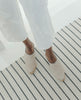 Top view shot of model on carpet wearing the Coclico Kera Shearling Clog in Latte nubuck, a slip-on mule with a mid-height wood block base, tapered toe, shearling lining. 4