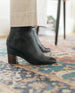 Model standing in Coclico Lono Boot in Prismanet Black leather, a textured leather wrapped demi-wedge boot with an inside zip closure, leather sole and a mid-height, gently curved solid wood heel - side view.  2