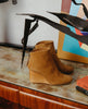 Side view of the Coclico Lovage Boot in Wheat suede: Sculpted 75mm solid wood wedge with a rounded toe and a back zip closure - placed on wood/marble table with decor in background. 4