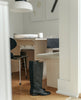 The Coclico Mine Boot in Black leather displayed in side view on wooden floor near dining nook. 2