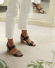 Woman modeling the Coclico Bita Heel in Deep Sea leather: Open sandal two straps across foot, elasticated back strap, gently curved wood block mid-height heel. 6