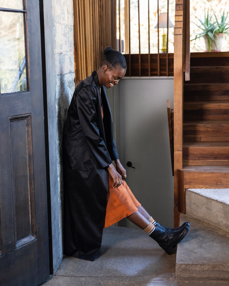 Woman smiling in glossy orange dress and black coat while leaning against a cement wall and wearing the Damiana Shearling Boot.