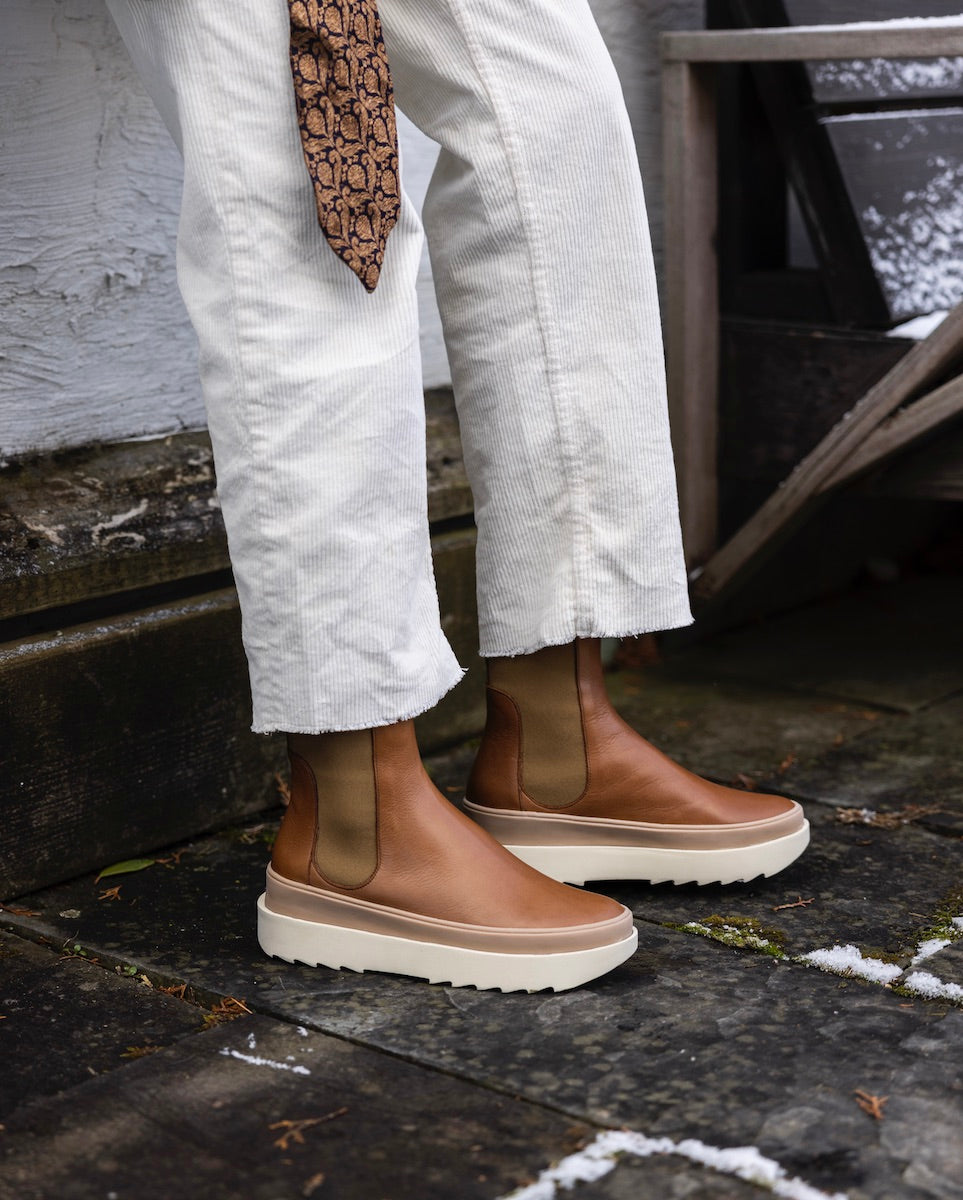 A pair of brown leather Chelsea boots with elastic side panels, white rubber soles, and a sleek, smooth texture, worn with white corduroy pants.