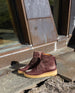 Sunny side view of the Hop Boot in Merlot leather placed outside in front of an industrial building.  2