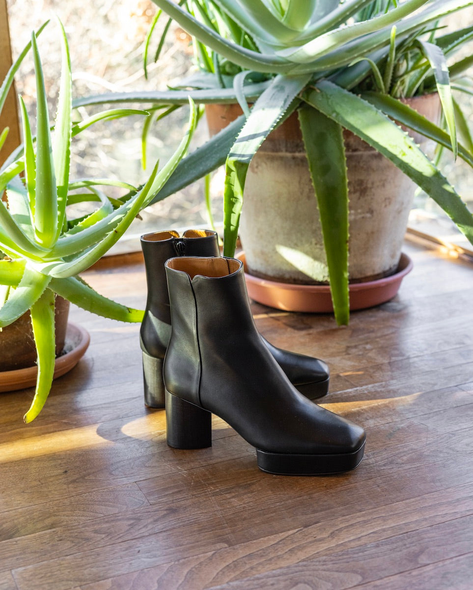 The Travis Boot in Black placed on a wooden floor with plants in the background.