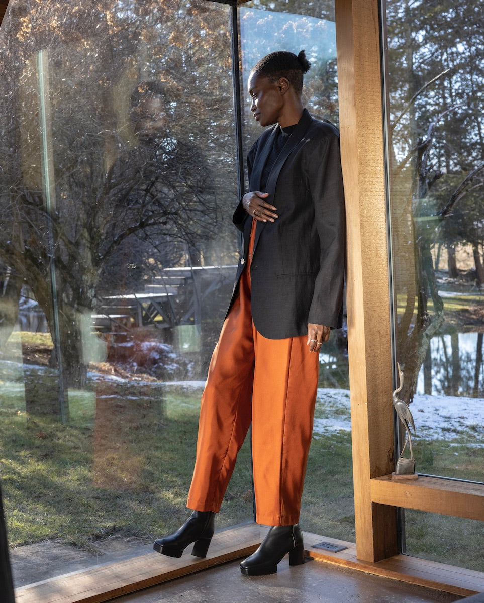 Model standing, leaning against the wall and wearing orange pants, a black blazer with the Travis Boot in Black.