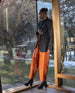 Model standing, leaning against the wall and wearing orange pants, a black blazer with the Travis Boot in Black.  6