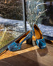 Trinity Pump in Painter's Blue in front of a glass window on a wooden floor.  2