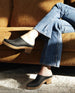 Woman seated cross legged modeling the Coclico Kule Clog in Black leather, a slip-on mule, tapered toe, mid-height solid wood base. 5