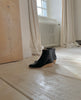 Side left view of the Java Boot in Black in a room with white flooring and a white radiator and curtains in the back. 6