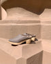 Coclico Kera Shearling Clog in Fog nubuck, a slip-on mule with a mid-height wood block base, tapered toe, shearling lining - placed on terracotta wall, outdoor shot.  4