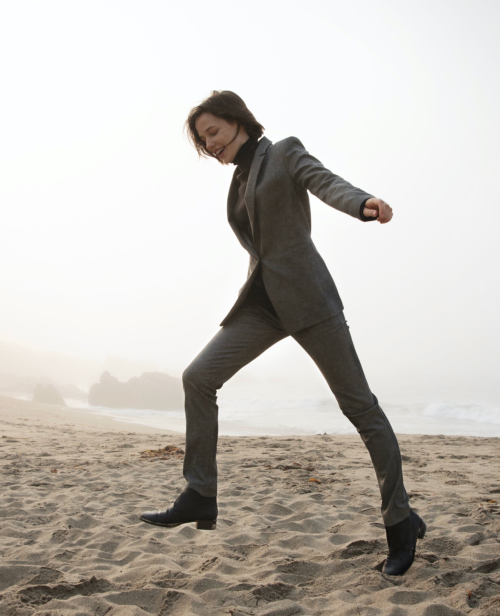 Woman wearing a black turtleneck with a grey suit and the Midori Bootie in Black skipping on a beach.