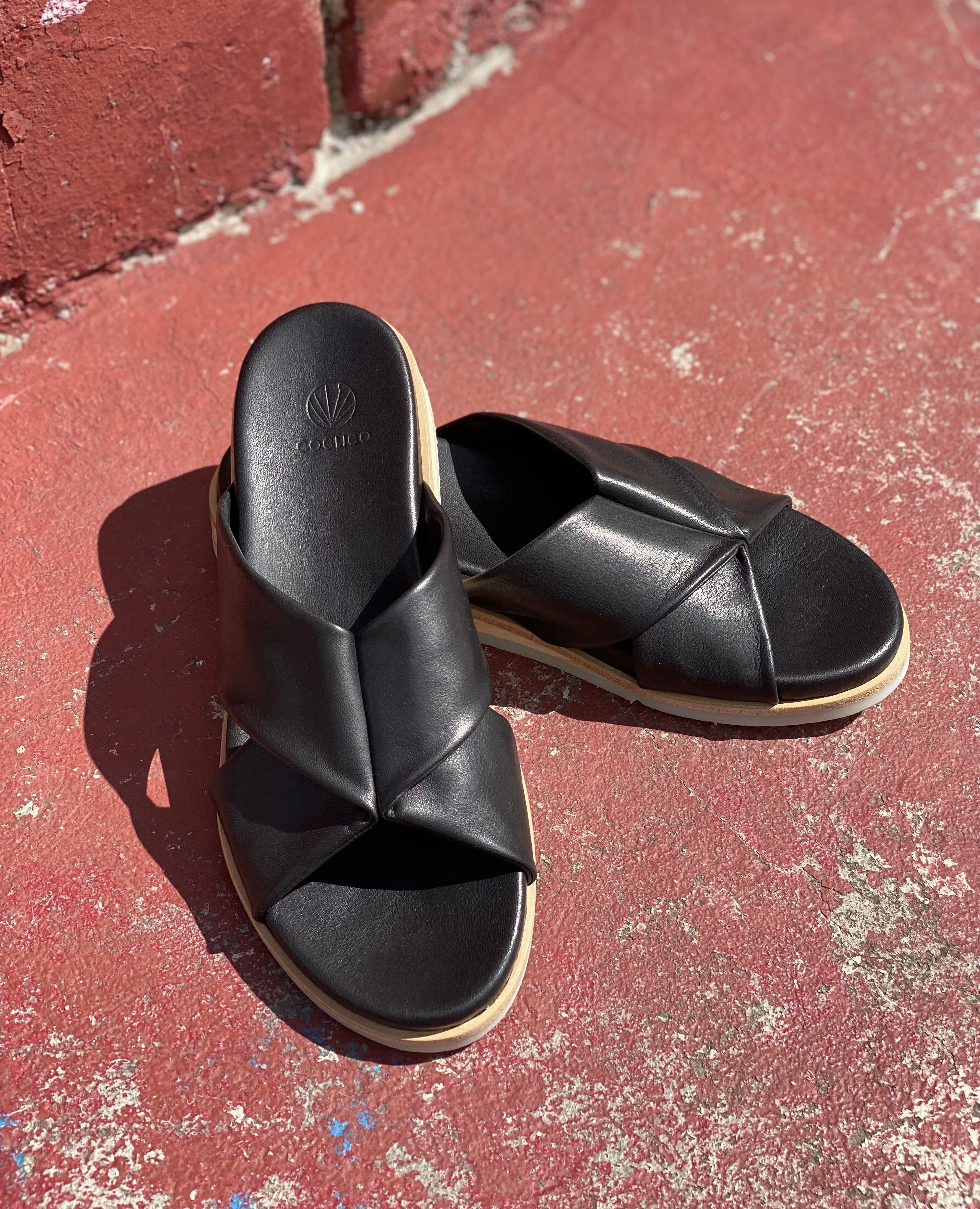Close up of Coclico Pasque Sandal in Black leather, a low pine wedge slide sandal with rubber sole and intertwined leather tube straps across foot - on red cement flooring in sunlight.