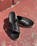 Close up of Coclico Pasque Sandal in Black leather, a low pine wedge slide sandal with rubber sole and intertwined leather tube straps across foot - on red cement flooring in sunlight.  8