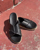 Close up of Coclico Pasque Sandal in Black leather, a low pine wedge slide sandal with rubber sole and intertwined leather tube straps across foot - on red cement flooring in sunlight. 8