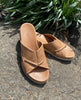 Close up of Coclico Pasque Sandal in Mandorla leather: a low pine wedge slide sandal, intertwined leather tube straps across foot, gum-rubber sole - on cement with plants in background. 7