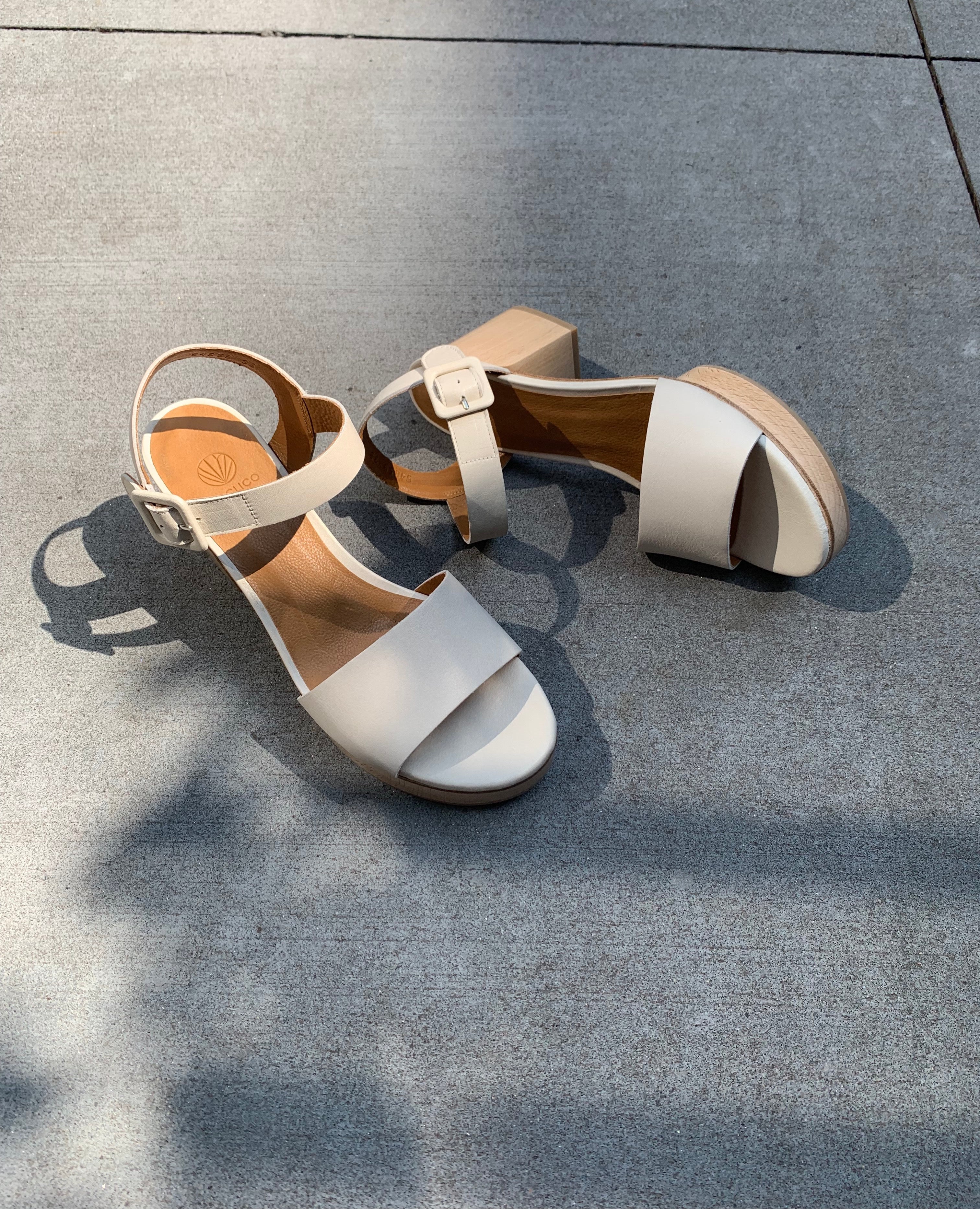Close up, sunlit shot of Coclico Riviera Clog in Greige (off-white) leather: wide front band, quarter-strap, buckle closure, with a solid wood platform to match the solid wood mid-height heel.