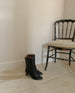 Wakame Boot in Black Leather placed next to a chair against a white wall.  2