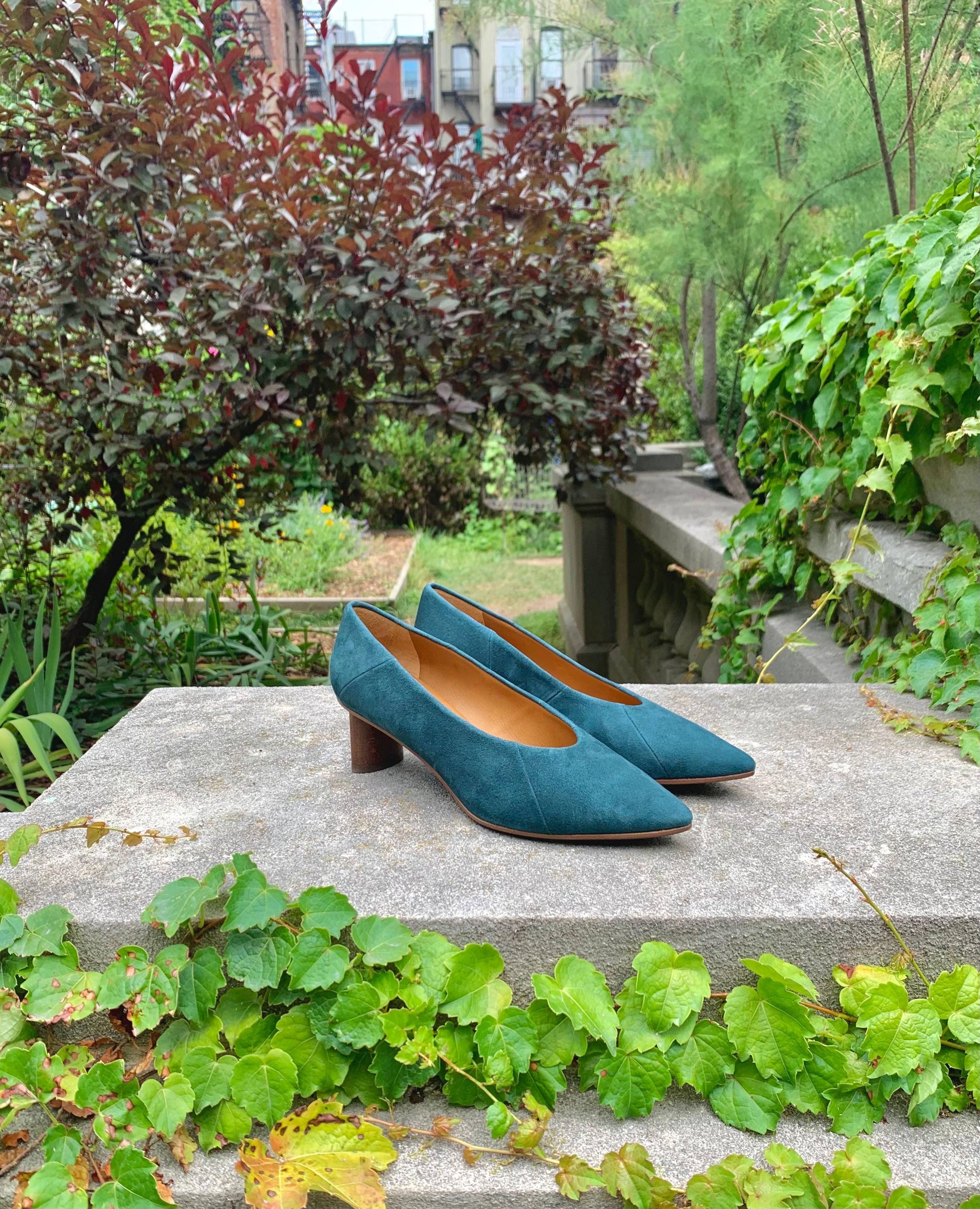The Wumo Heel in Bottle suede on a stone block in flourishing garden.