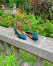 Coclico Wumo Heel in Bottle suede with a scultped, oval shaped solid wood heel in a hand-painted chestnut finish - placed on stone wall with greenery in background. 5