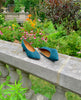 Coclico Wumo Heel in Bottle suede with a scultped, oval shaped solid wood heel in a hand-painted chestnut finish - placed on stone wall with greenery in background. 5