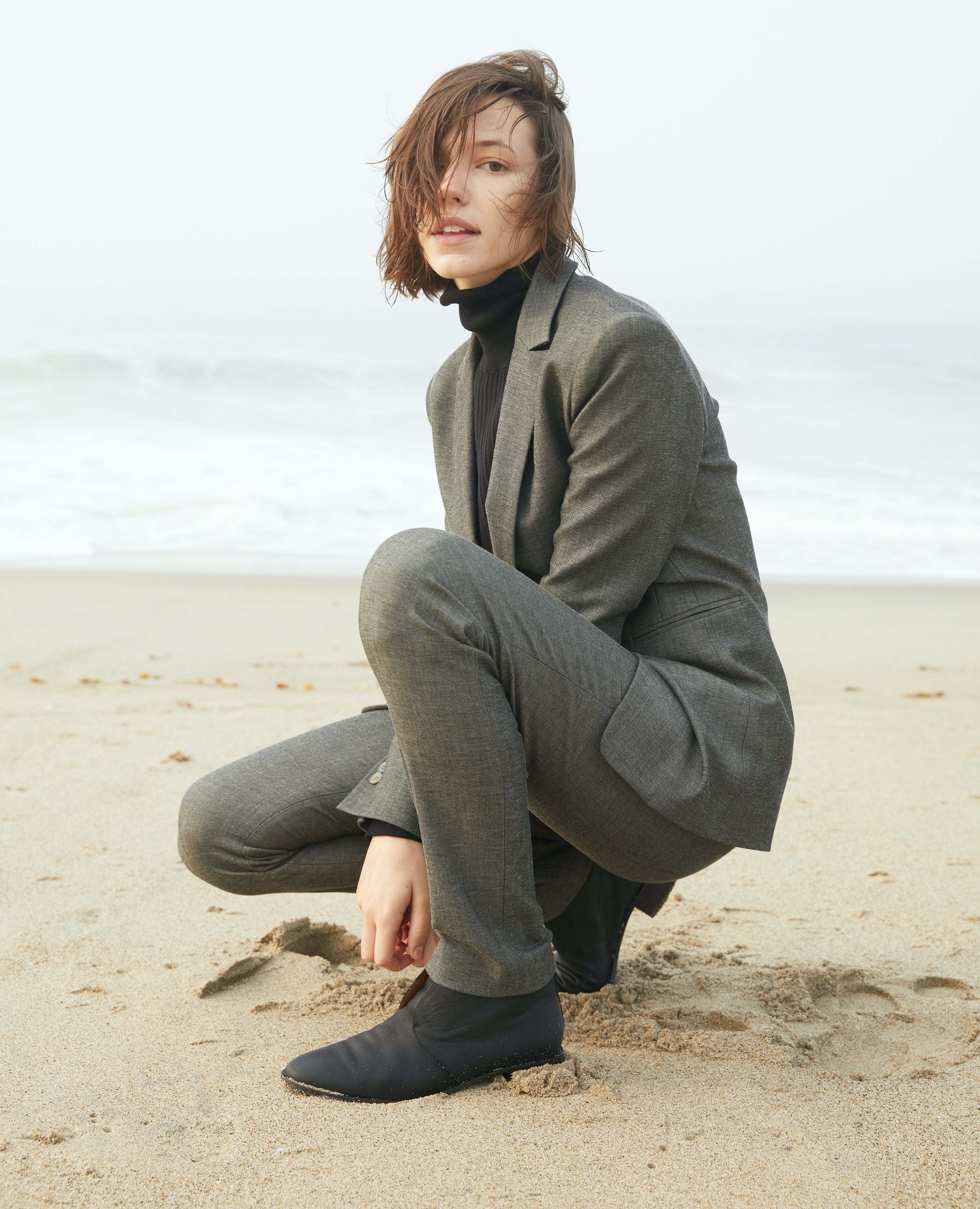 Woman wearing a black turtleneck, grey suit and Midori Bootie crouched down in the sand on a beach.