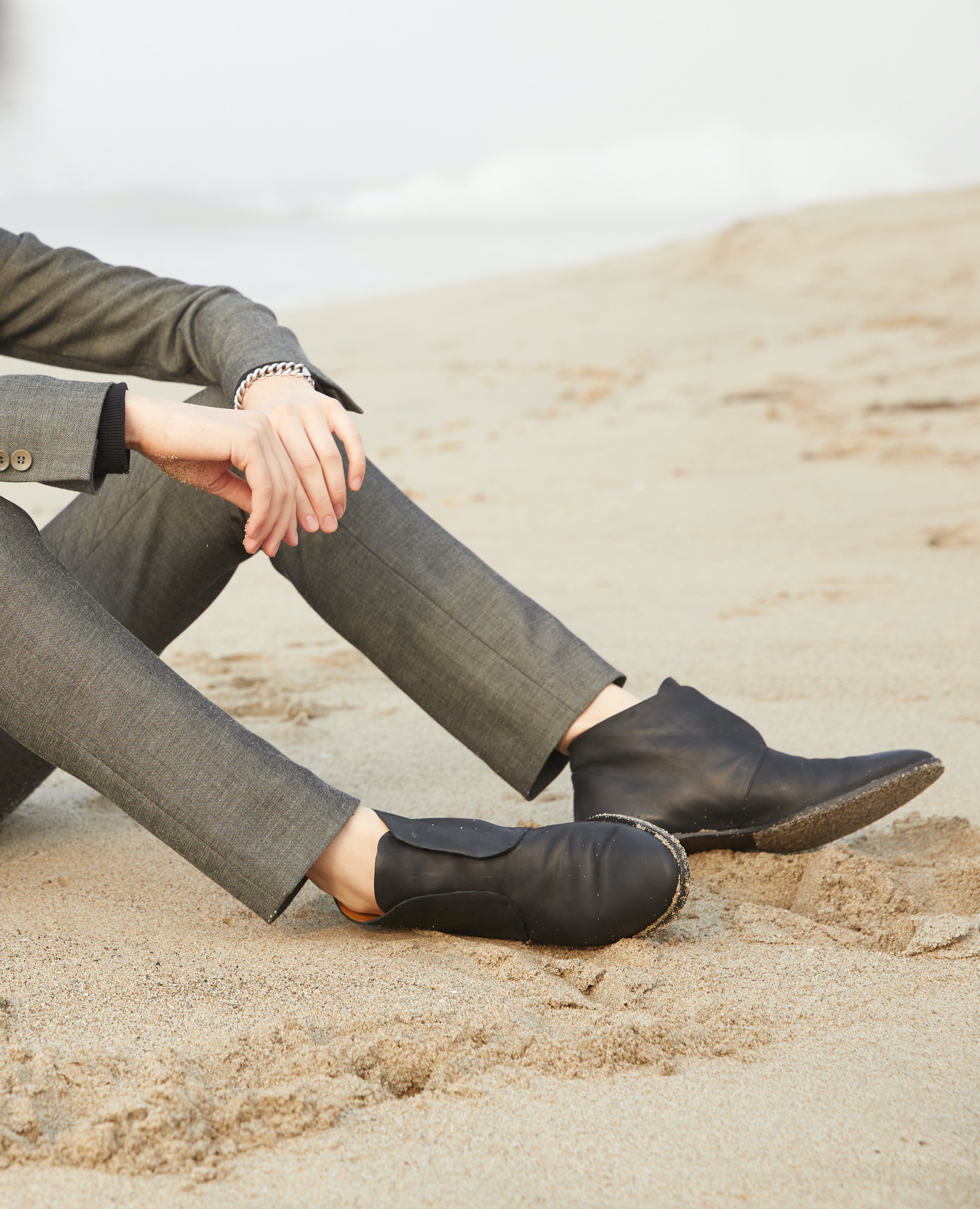 Hands, legs, and feet of a woman sitting on the beach wearing a suit and the Midori Boot in Black.