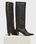 Tall, black leather boots with a sleek, smooth texture, pointed toes, mid-height heels, and side zippers, standing on a light-colored textured pedestal. 3