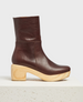 A mid-calf, dark brown leather clog boot with a smooth texture, featuring a chunky, light wooden heel and platform, displayed on a textured, off-white pedestal. 1