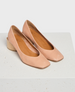 A pair of elegant, blush pink leather flats with a smooth texture, featuring a low, chunky wooden heel and a rounded square toe design. 1