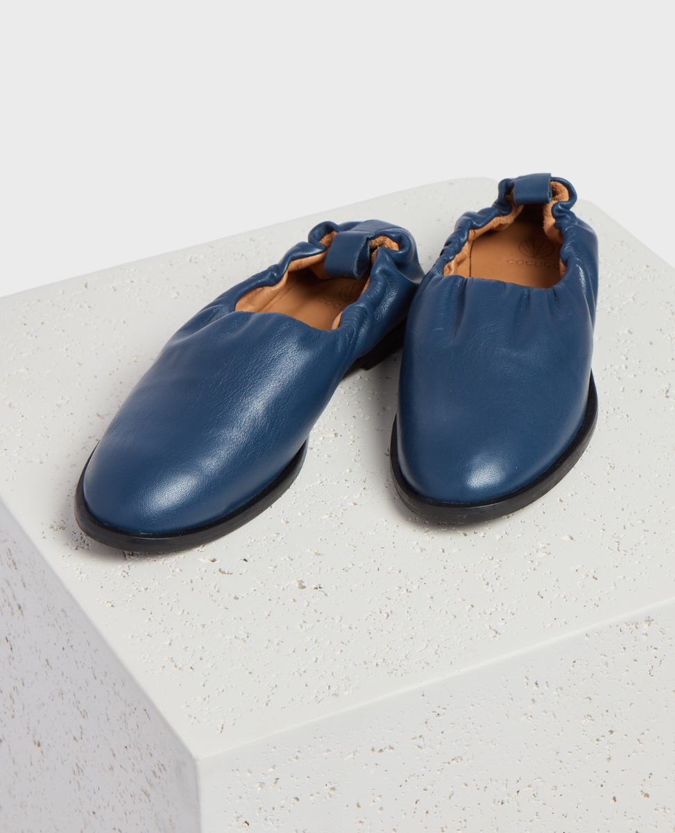A pair of blue leather slip-on shoes with a smooth texture, rounded toe, elasticized back, and black soles, displayed on a textured white surface.