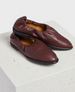 A pair of burgundy leather slip-on shoes with a soft, gathered texture, featuring a rounded toe and a black sole, resting on a textured white surface. 1