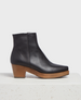 A sleek, black leather ankle boot with a smooth texture, featuring a square toe, side zipper, and a contrasting wooden block heel. 1