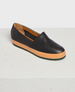 A sleek, black leather slip-on shoe with a smooth texture, featuring a contrasting tan rubber sole and a minimalist design. 1